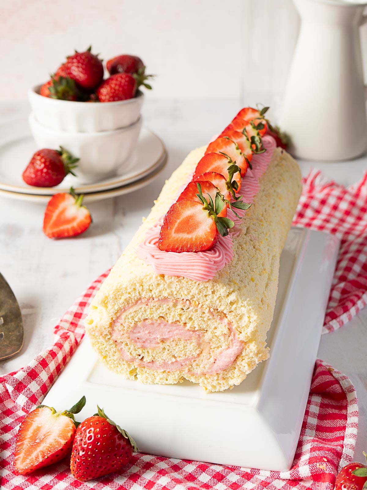 A strawberry Swiss roll cake with pink cream filling and sliced strawberries on top, placed on a white platter next to fresh strawberries and a red checkered cloth.