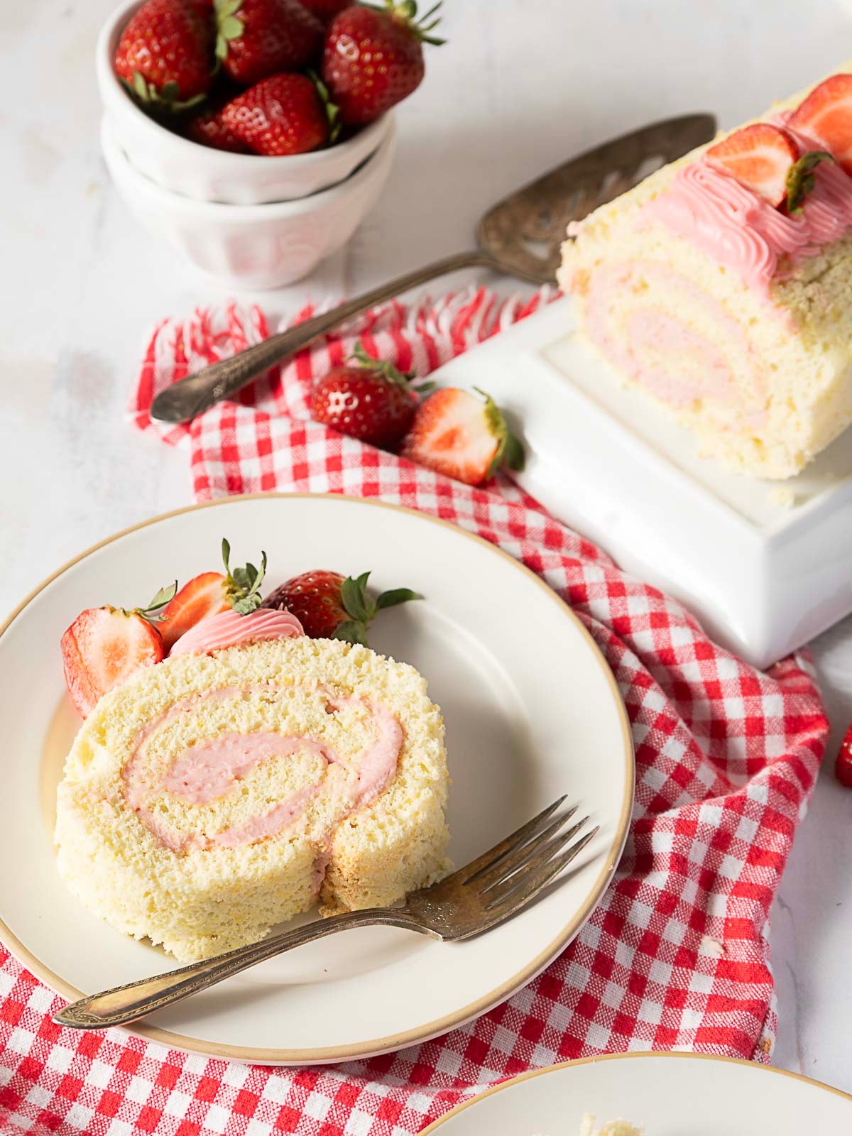 A slice of strawberry Swiss roll cake on a plate with a fork, garnished with fresh strawberries; the rest of the cake and more strawberries are in the background.