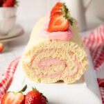 A rolled sponge cake filled and topped with pink strawberry cream, garnished with fresh strawberries, sits on a white plate with a red checked cloth underneath.