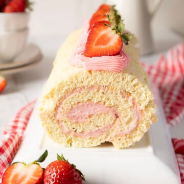 A rolled sponge cake filled and topped with pink strawberry cream, garnished with fresh strawberries, sits on a white plate with a red checked cloth underneath.