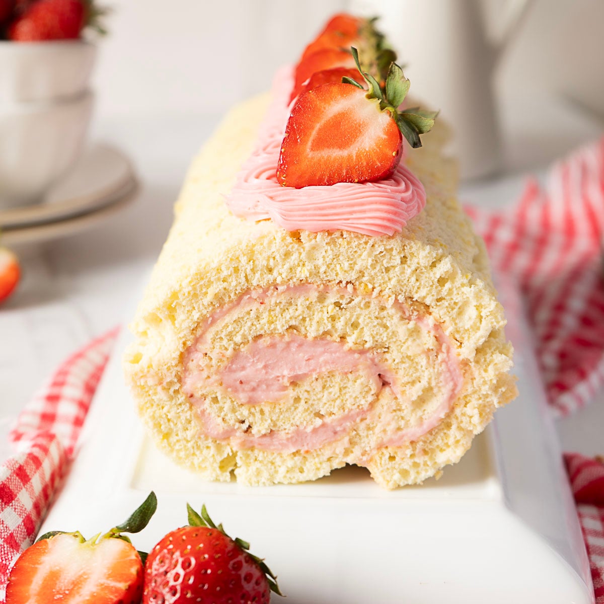 A rolled sponge cake filled and topped with pink strawberry cream, garnished with fresh strawberries, sits on a white plate with a red checked cloth underneath.