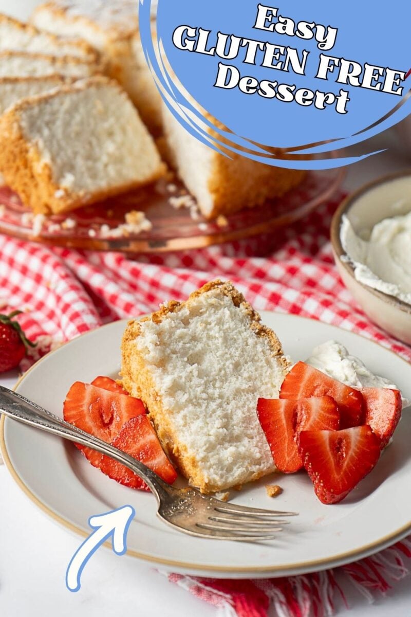 A plate with a slice of Gluten Free Angel Food Cake, fresh sliced strawberries, and whipped cream sits on a red checkered cloth. Whole cake and a bowl of whipped cream are in the background.