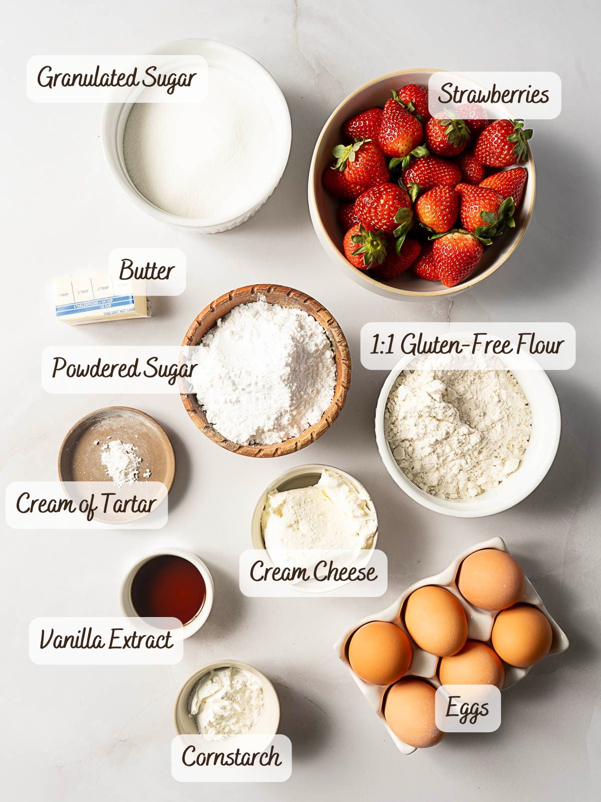 Bowls and containers of ingredients labeled: granulated sugar, strawberries, butter, powdered sugar, gluten-free flour, cream of tartar, cream cheese, vanilla extract, cornstarch, and eggs.