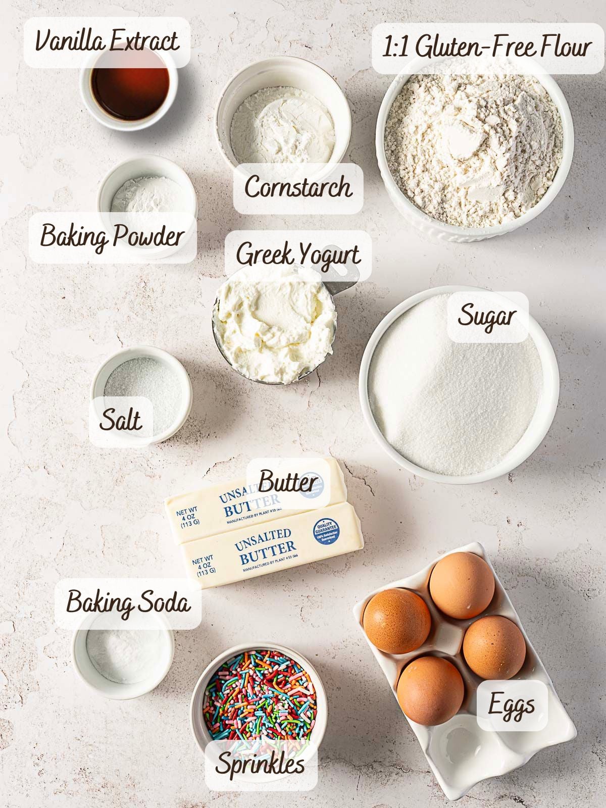 Top-down view of labeled baking ingredients for a gluten free birthday cake, including gluten-free flour, sugar, butter, eggs, Greek yogurt, baking powder, baking soda, cornstarch, salt, vanilla extract, and sprinkles.