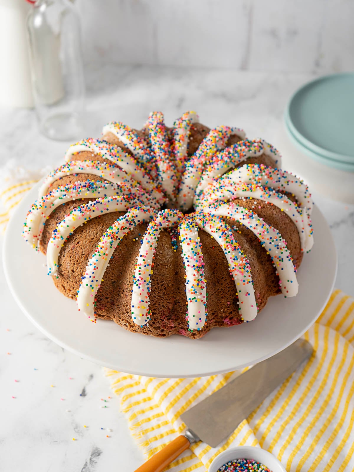 A bundt cake with white icing drizzled on top and colorful sprinkles, displayed on a white cake stand with a knife and yellow-striped towel nearby.