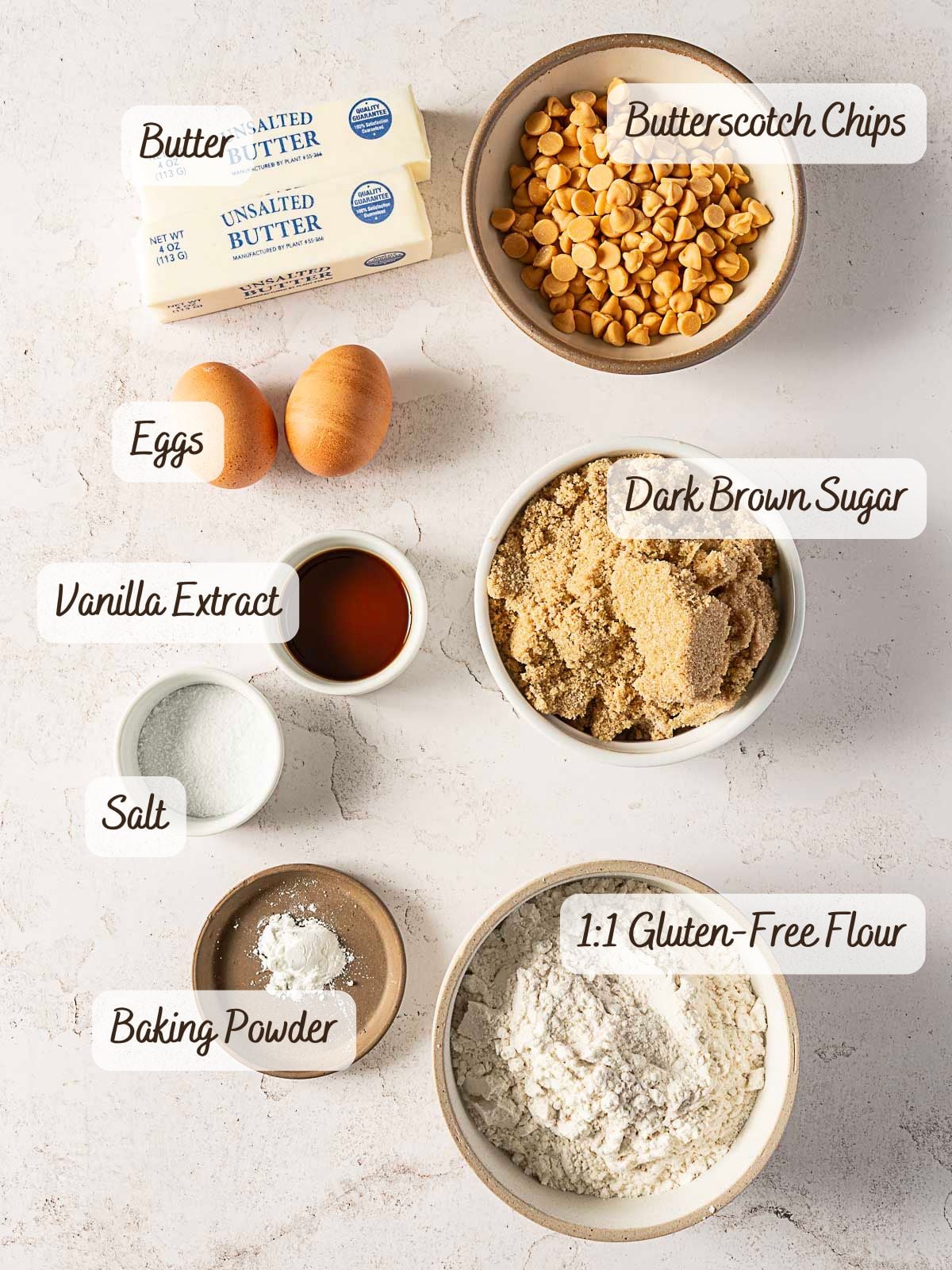 Top-down view of labeled baking ingredients: butter, eggs, vanilla extract, salt, baking powder, 1:1 gluten-free flour, dark brown sugar, and butterscotch chips on a light surface.