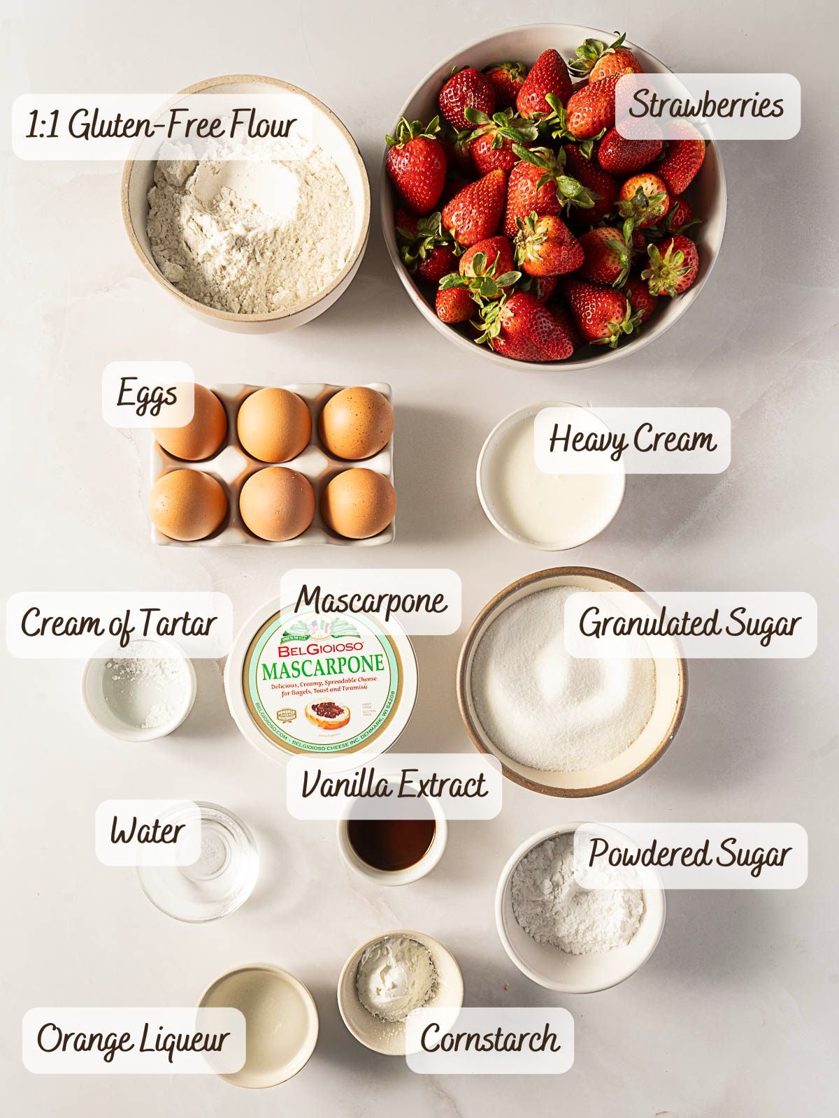 Various baking ingredients are arranged on a table, including strawberries, eggs, gluten-free flour, mascarpone, heavy cream, sugars, vanilla extract, cream of tartar, cornstarch, water, and orange liqueur.
