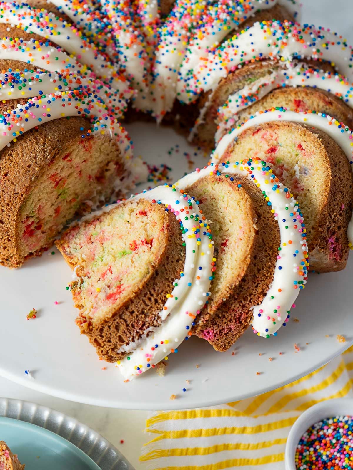 A Bundt cake with white icing and colorful sprinkles is sliced and arranged on a plate, with a few sprinkles scattered on the surface.