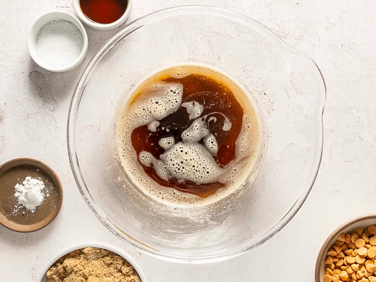 A glass mixing bowl with melted butter and vanilla, surrounded by small bowls of brown sugar, peanut butter chips, salt, and baking powder on a white countertop.
