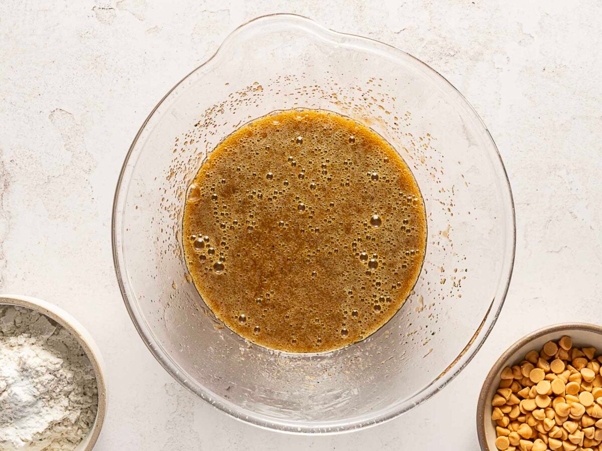 A glass mixing bowl containing a blended brown liquid mixture, with bowls of flour and butterscotch chips nearby on a light countertop.