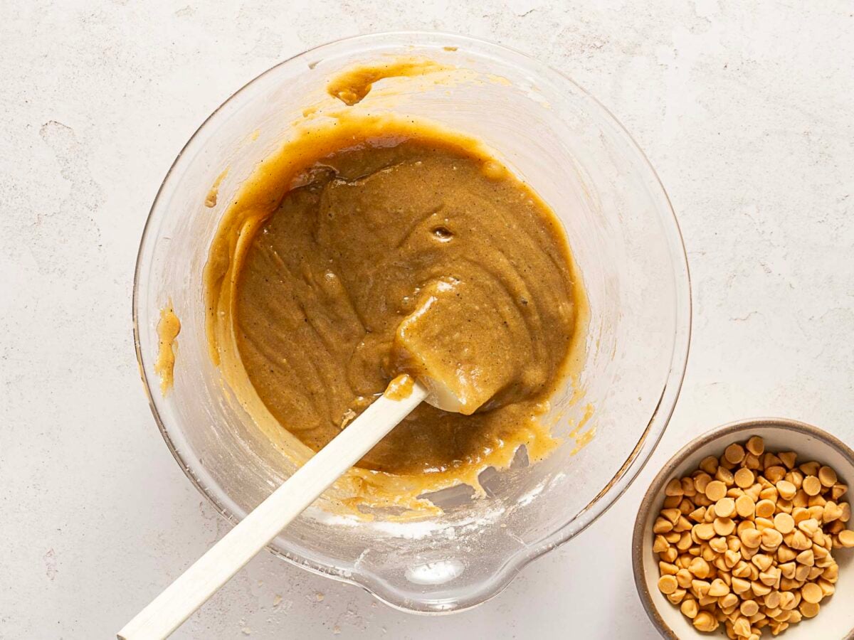 A glass mixing bowl with cookie dough and a white spatula next to a small bowl of butterscotch chips on a light surface.