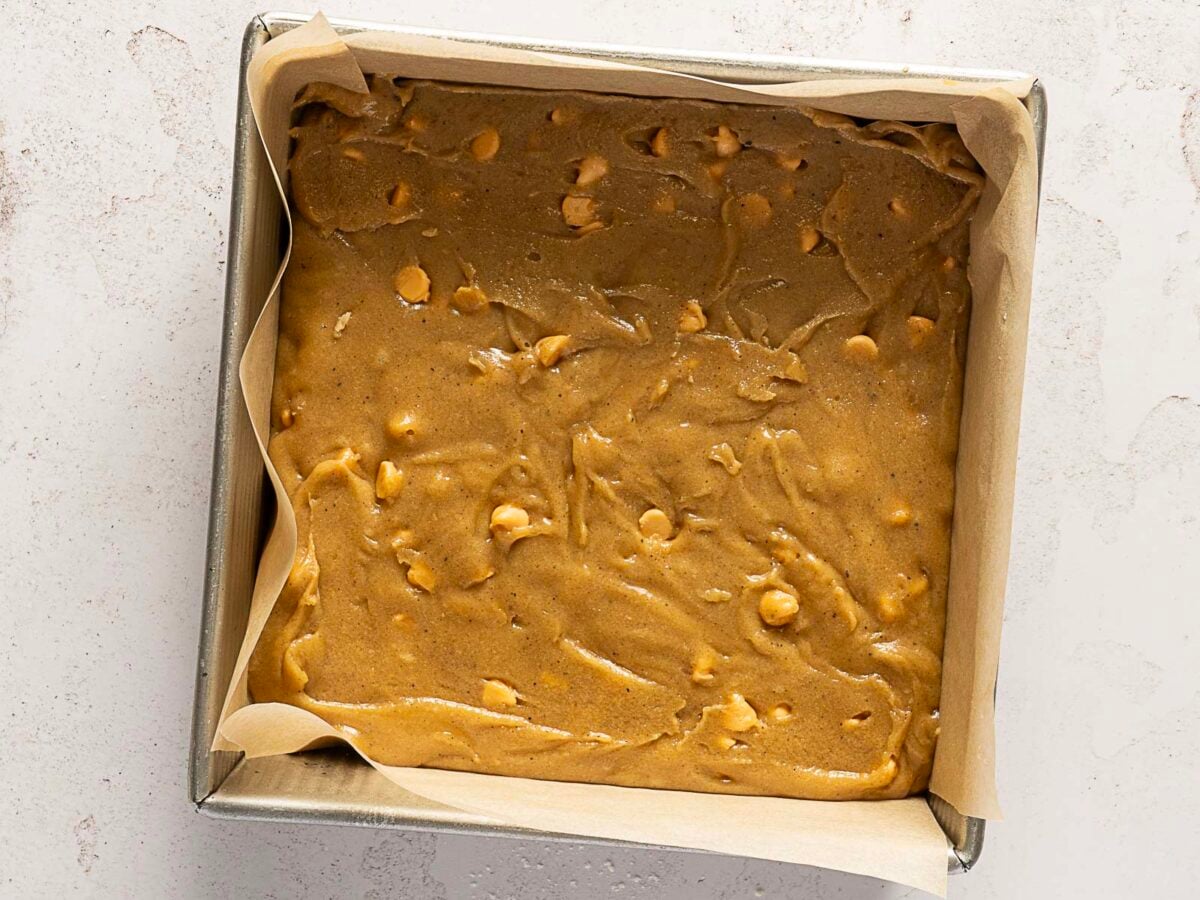 A square baking pan lined with parchment paper is filled with unbaked, light brown cookie dough containing white chocolate chips.