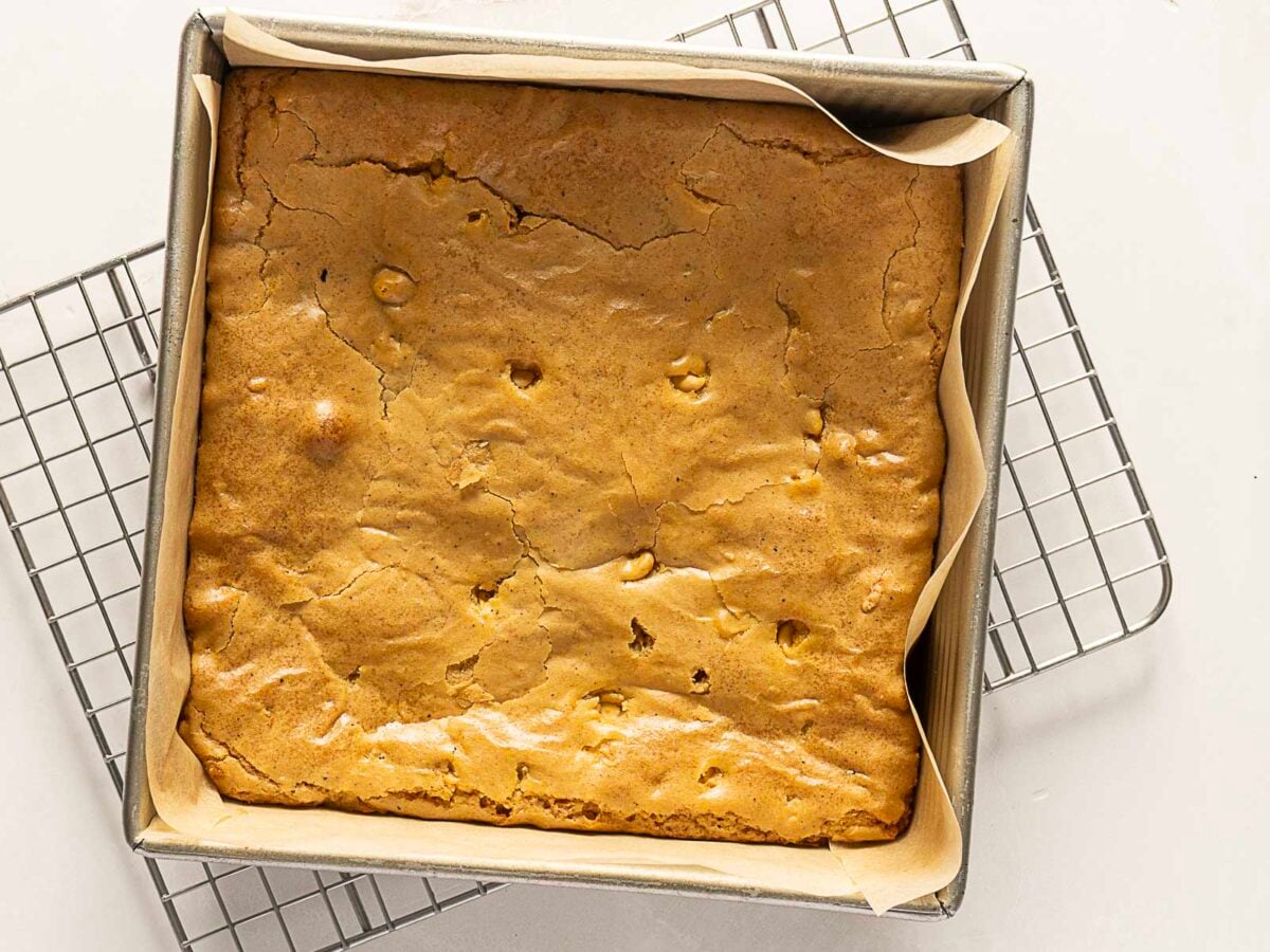 A square pan lined with parchment paper holds a freshly baked golden-brown blondie, resting on a metal cooling rack.
