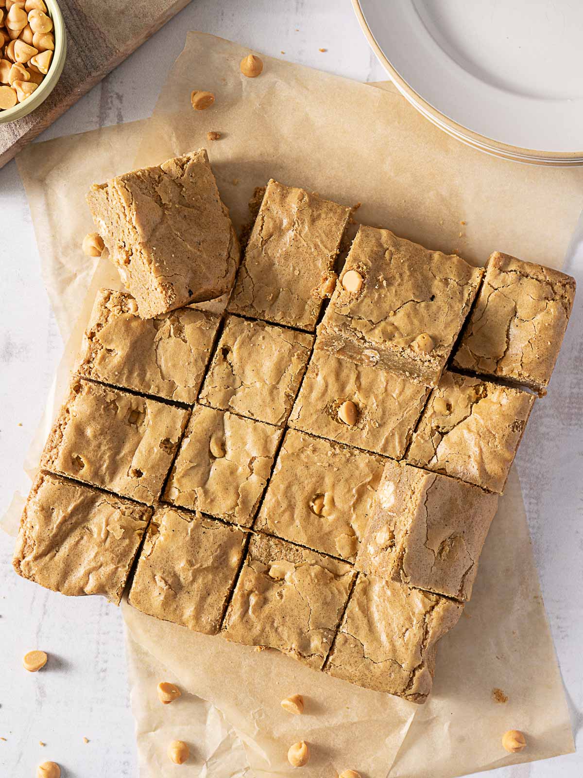 A batch of blondies cut into sixteen squares rests on parchment paper, with butterscotch chips scattered nearby.