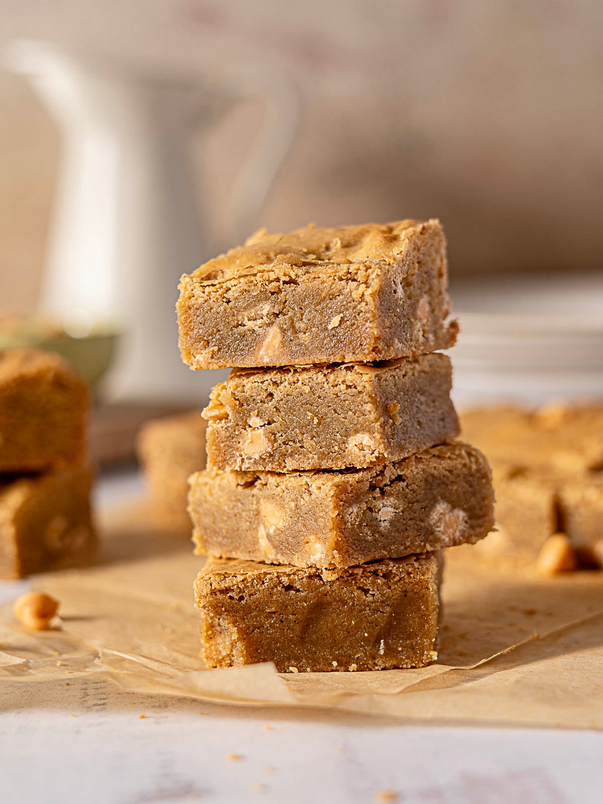 A stack of four peanut butter blondies sits on parchment paper, with more blondies and a white pitcher blurred in the background.