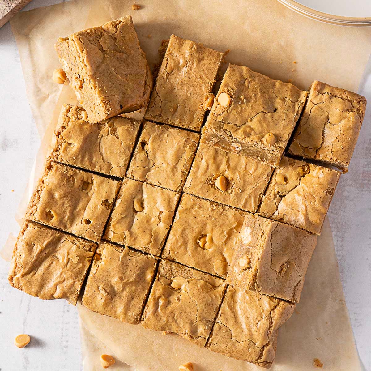 A batch of blondies cut into 16 square pieces, some pieces slightly separated, with visible butterscotch chips on top and parchment paper underneath.