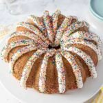 A bundt cake topped with white icing drizzles and colorful sprinkles, displayed on a white plate.