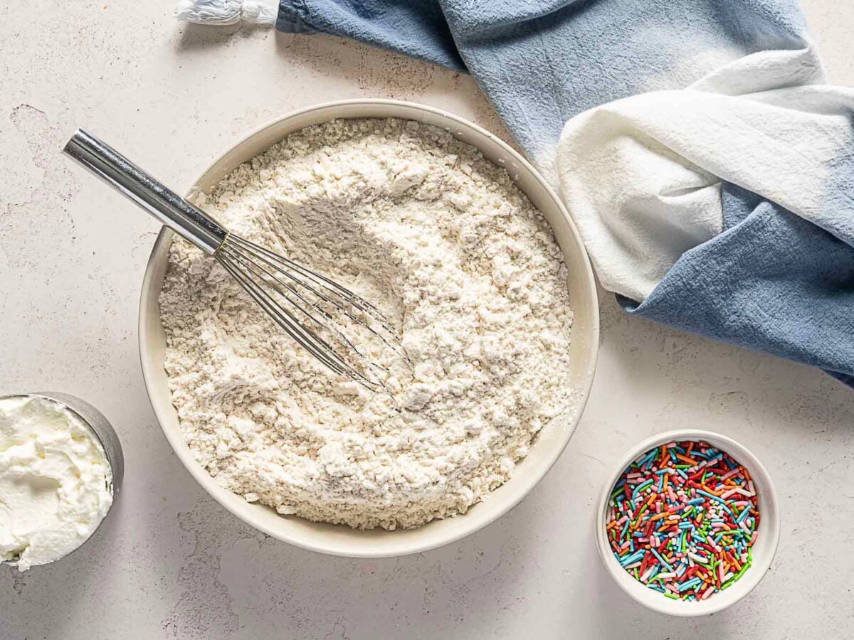 A bowl of flour with a whisk, a cup of white cream, and a bowl of colorful sprinkles on a light countertop next to a blue and white kitchen towel.
