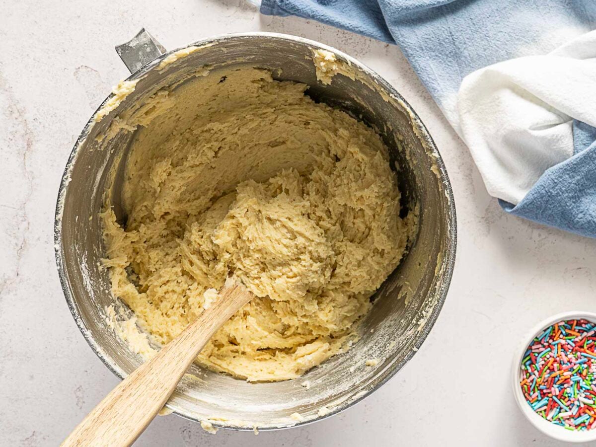 A mixing bowl filled with cookie dough and a wooden spoon, with a blue towel and a small bowl of colorful sprinkles nearby.