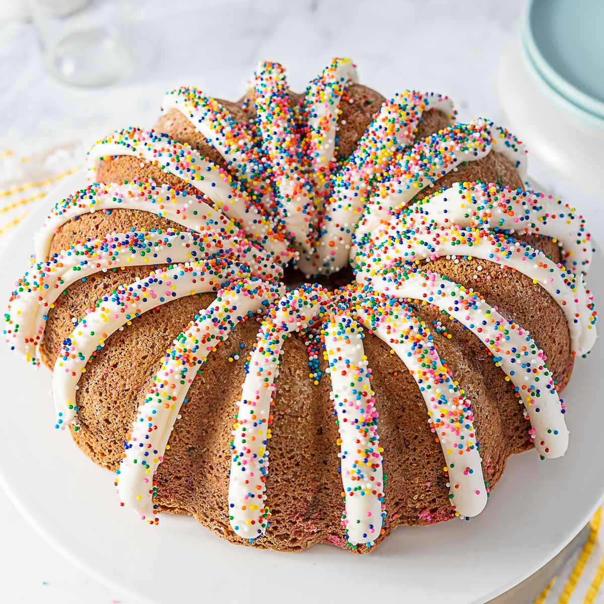 A bundt cake topped with white icing drizzles and colorful sprinkles, displayed on a white plate.