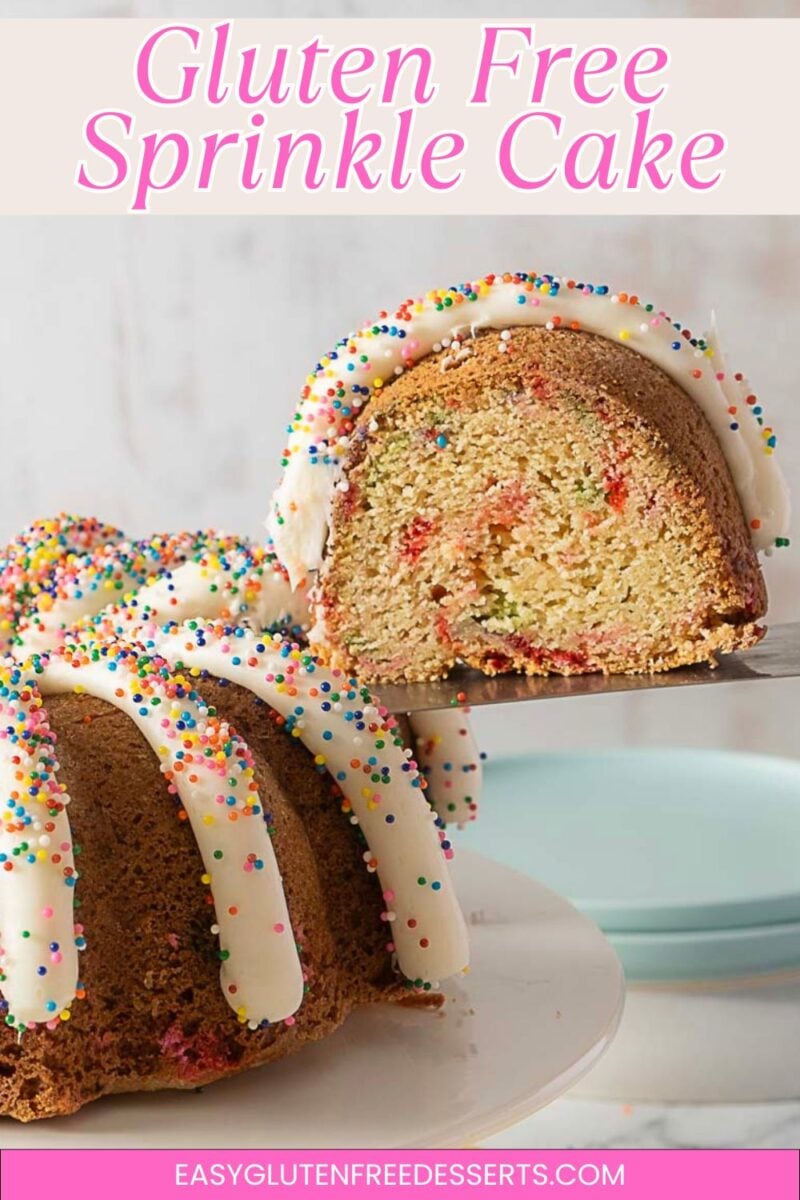 A gluten-free sprinkle bundt cake with white icing and colorful sprinkles, partially sliced, on a white cake stand with stacked plates in the background.