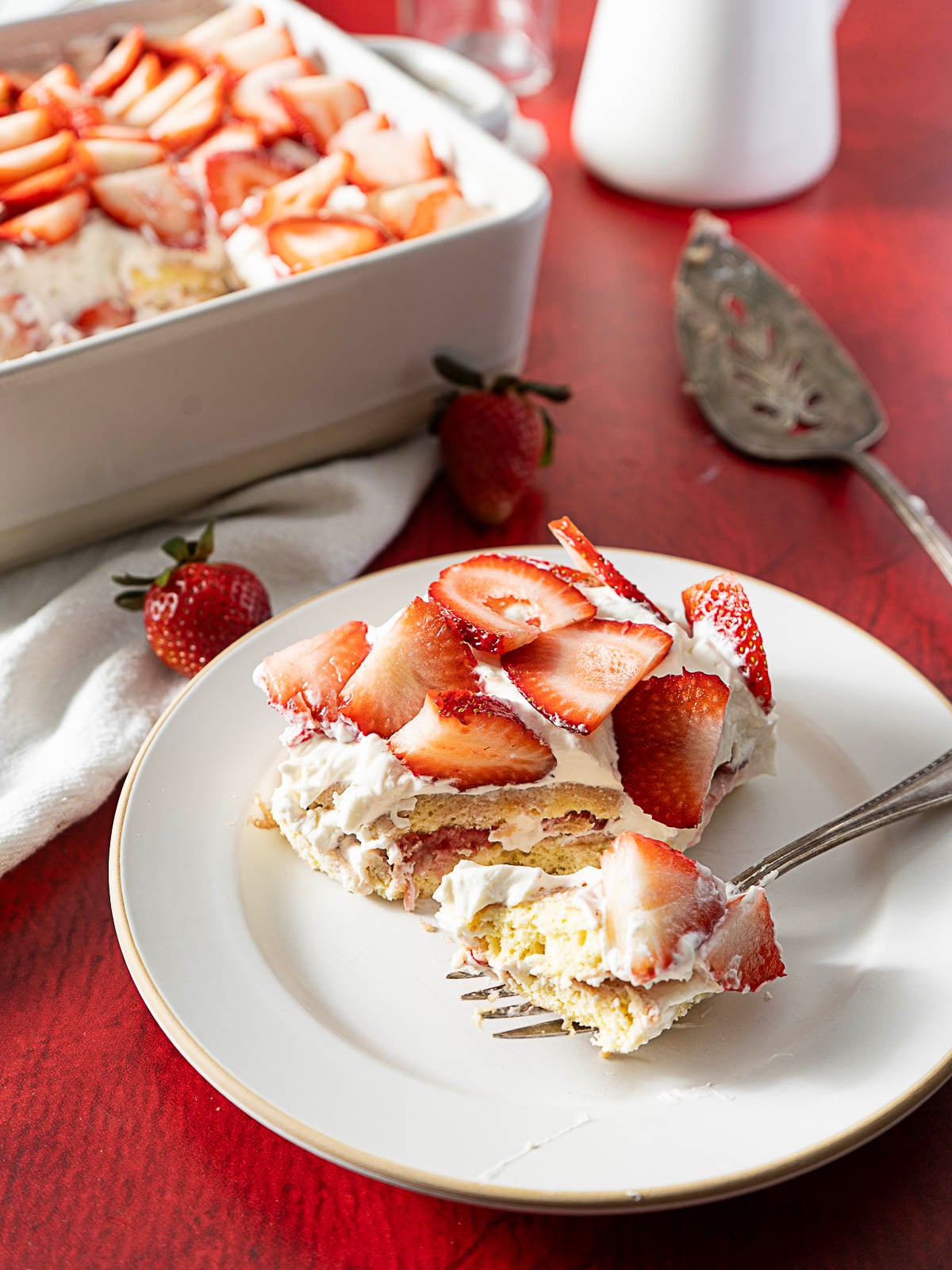 A slice of strawberry shortcake with whipped cream on a plate, topped with fresh strawberry slices, with a fork and whole strawberries nearby.