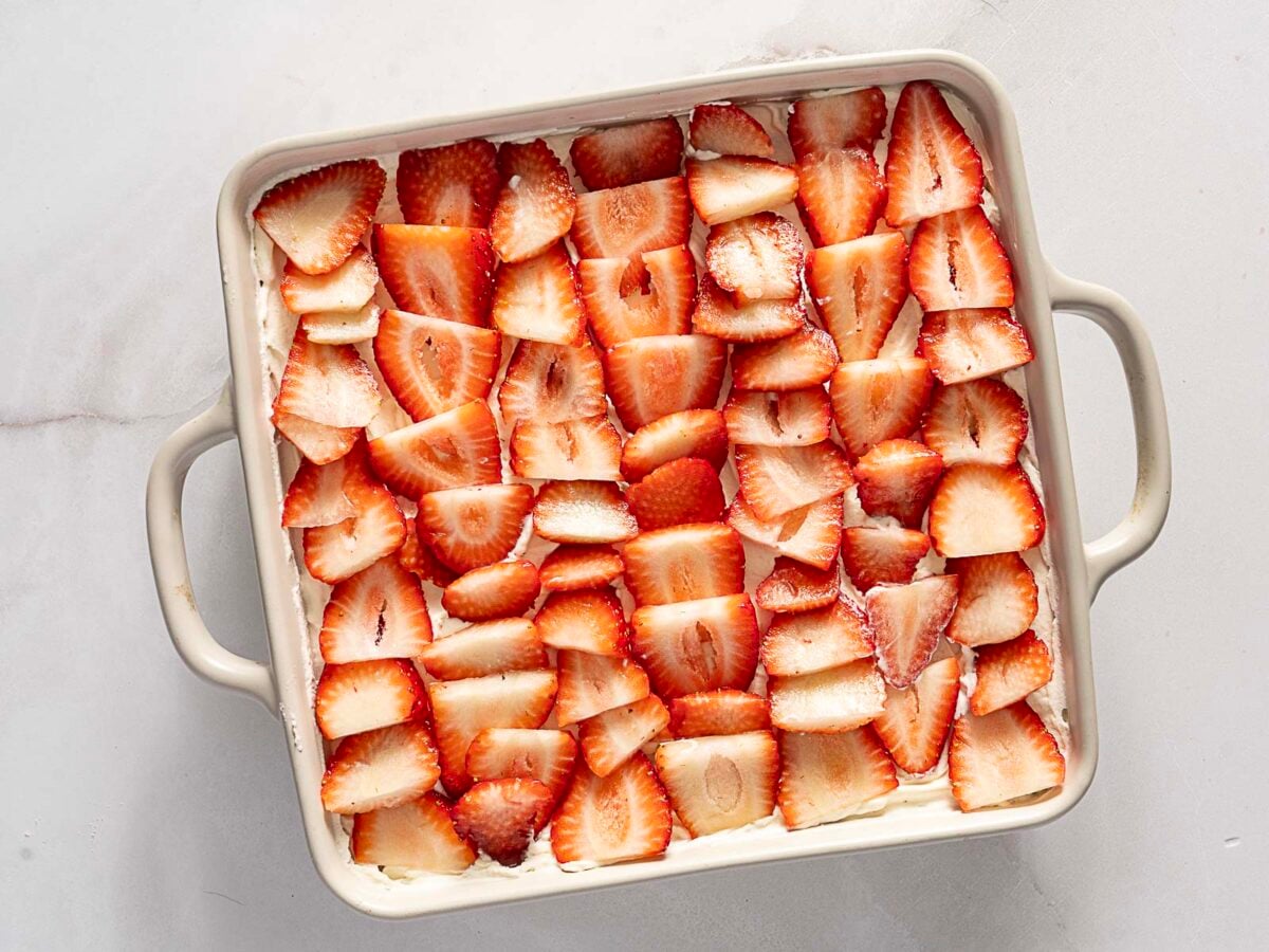 A square ceramic baking dish filled with sliced strawberries arranged in a single layer over a creamy base.