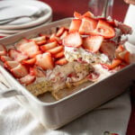 A square baking dish with a strawberry shortcake dessert; a spatula lifts a slice topped with sliced strawberries and cream. Plates and utensils are in the background.