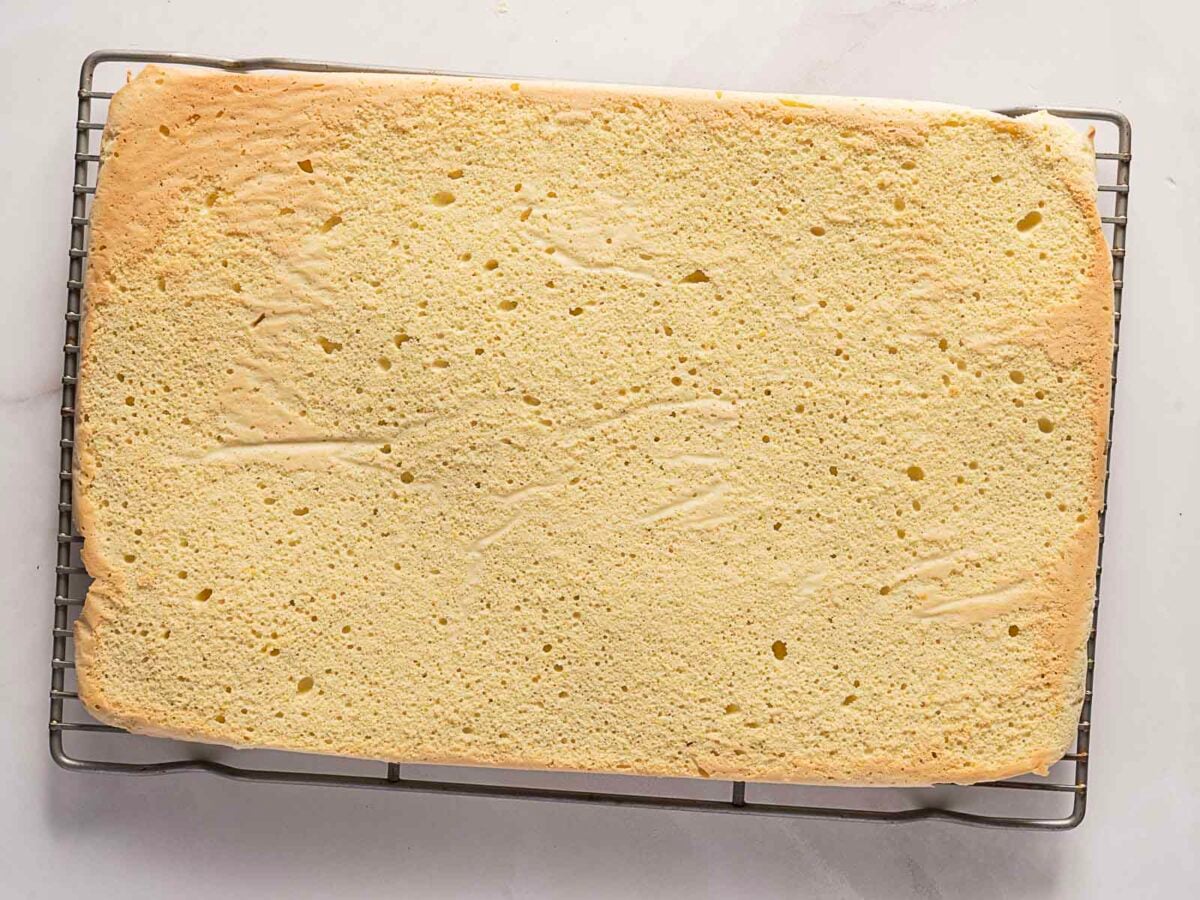 A rectangular sheet of sponge cake cooling on a metal wire rack, viewed from above on a white surface.