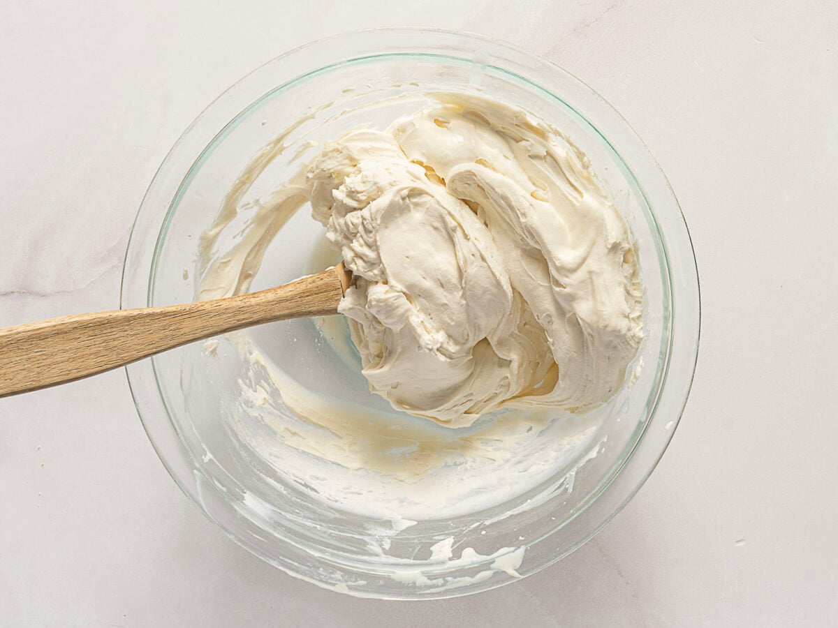 A glass bowl containing thick, creamy mixture being stirred with a wooden spatula on a light surface.