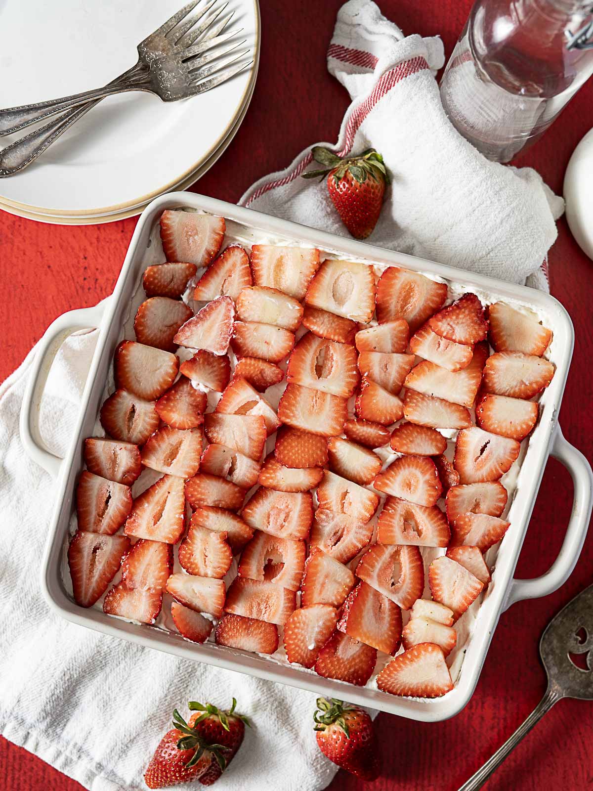 A square white baking dish filled with neatly arranged sliced strawberries, placed on a white towel with whole strawberries, plates, and cutlery nearby.