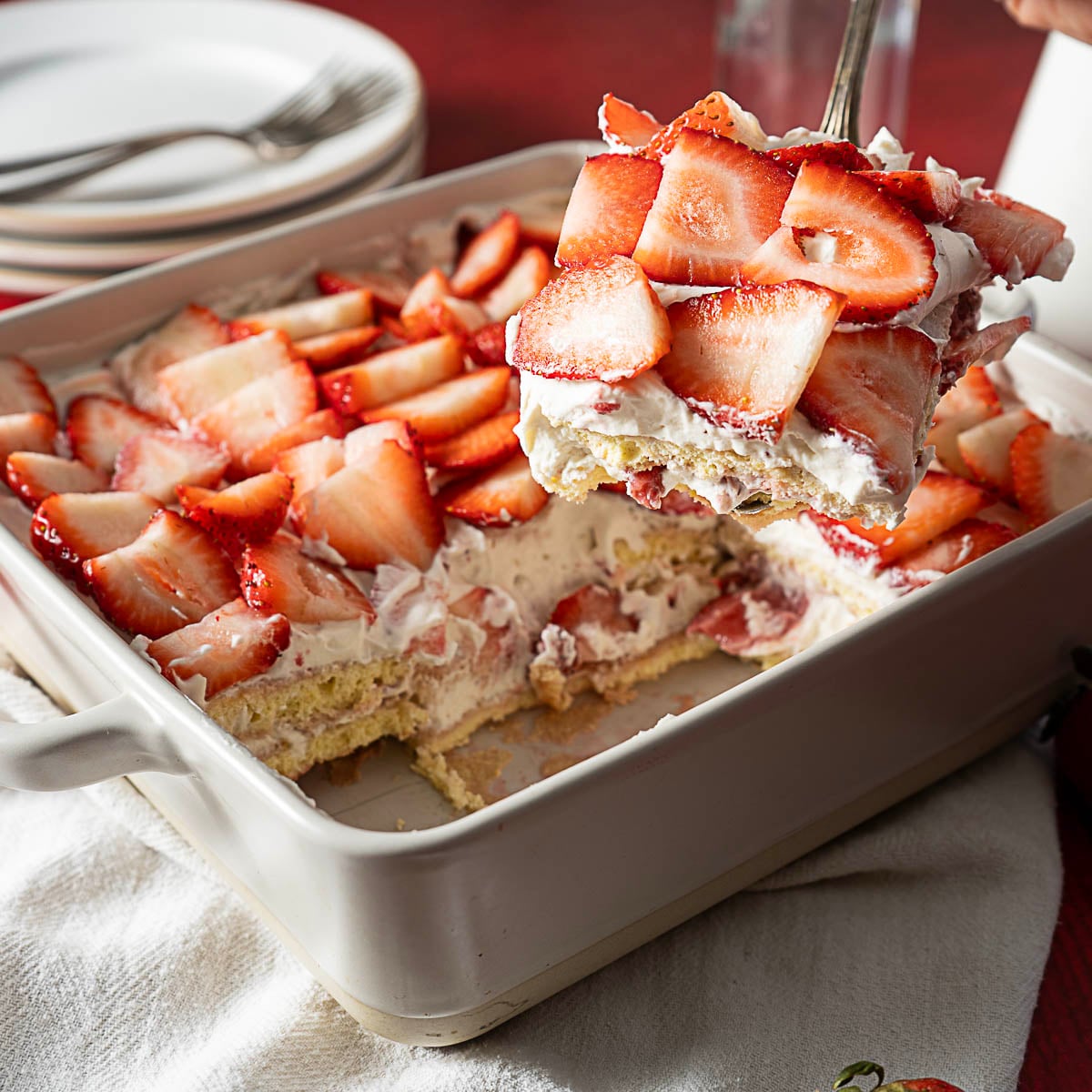 A square baking dish with a strawberry shortcake dessert; a spatula lifts a slice topped with sliced strawberries and cream. Plates and utensils are in the background.