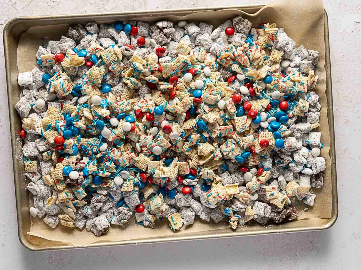A baking sheet lined with parchment paper holds a mix of Chex cereal, powdered sugar, red, white, and blue candies, and colorful sprinkles.