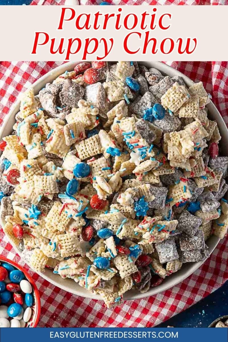 A bowl of patriotic puppy chow snack mix with Chex cereal, coated in powdered sugar and chocolate, mixed with red, white, and blue candies, on a red checkered cloth.