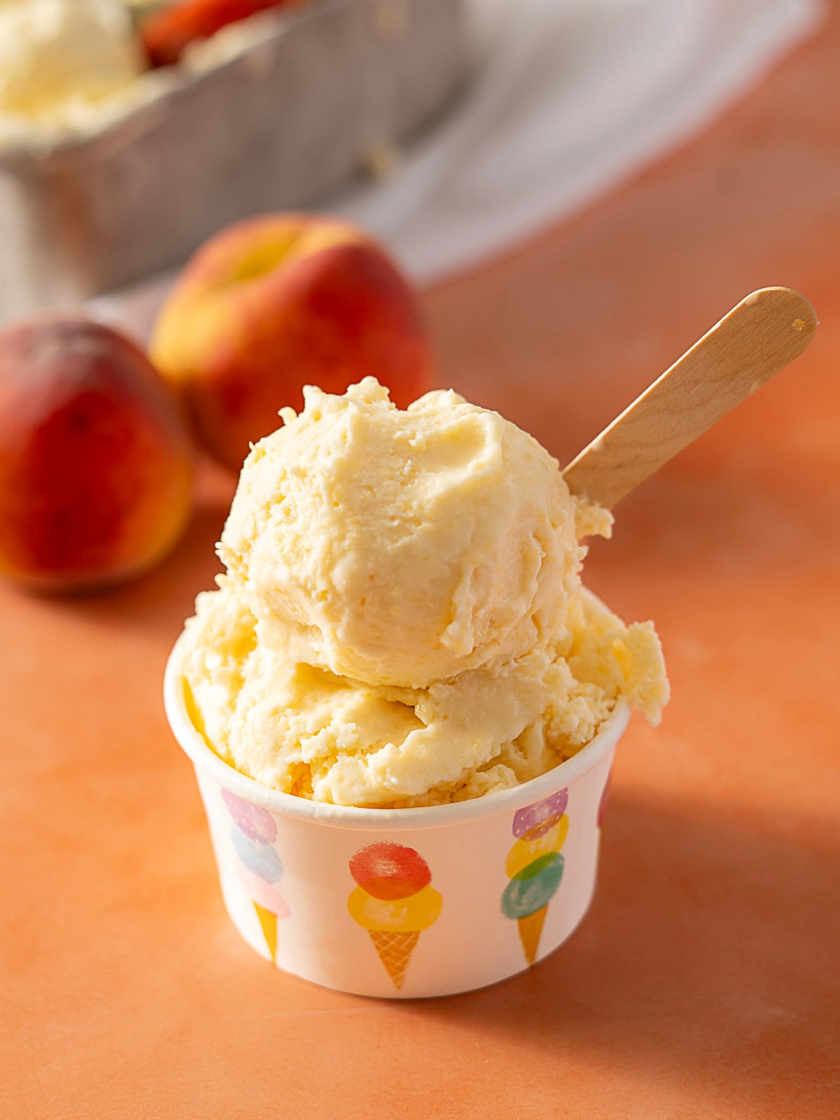 A cup of peach ice cream with a wooden spoon, with two peaches and a container of ice cream in the background on an orange surface.