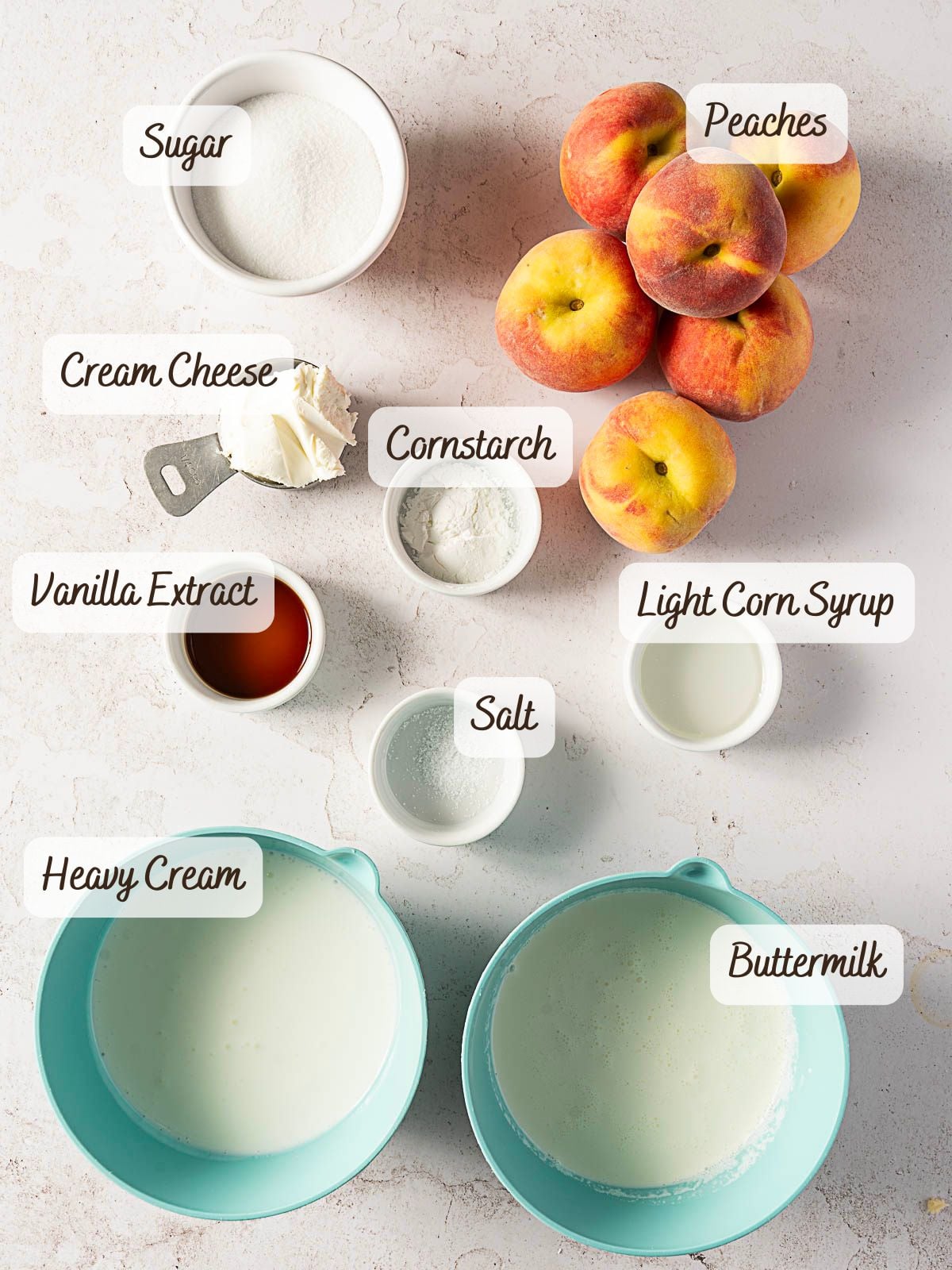 Labeled ingredients for a recipe arranged on a white surface, including sugar, peaches, cream cheese, cornstarch, vanilla extract, light corn syrup, salt, heavy cream, and buttermilk.