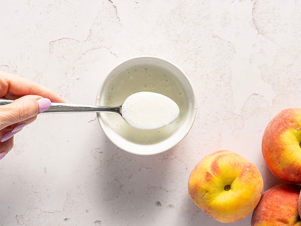 A hand holds a spoon of white yogurt above a bowl, with three peaches nearby on a light stone surface.