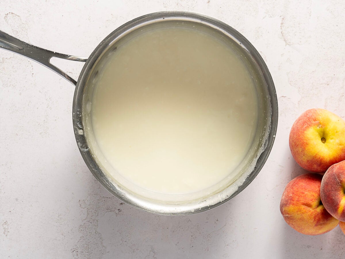 A saucepan filled with a creamy white mixture sits on a light surface next to three ripe peaches.