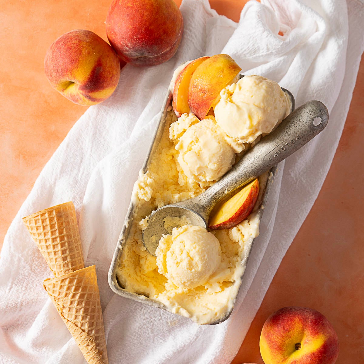 A metal container with peach ice cream scoops and a metal scoop, surrounded by fresh peaches and empty waffle cones on a white cloth.