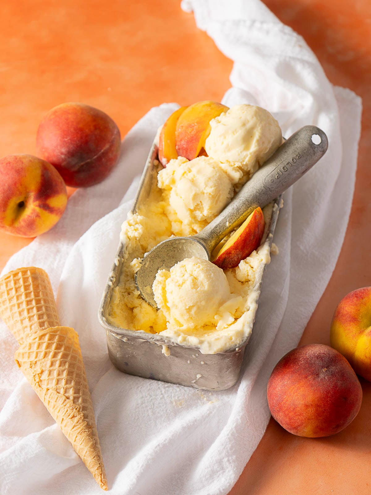 A metal container of peach ice cream with scoops and peach slices, surrounded by fresh peaches and waffle cones on a white cloth.