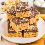 A plate of rice crispy treats topped with a layer of chocolate and chopped peanuts, stacked on top of each other.