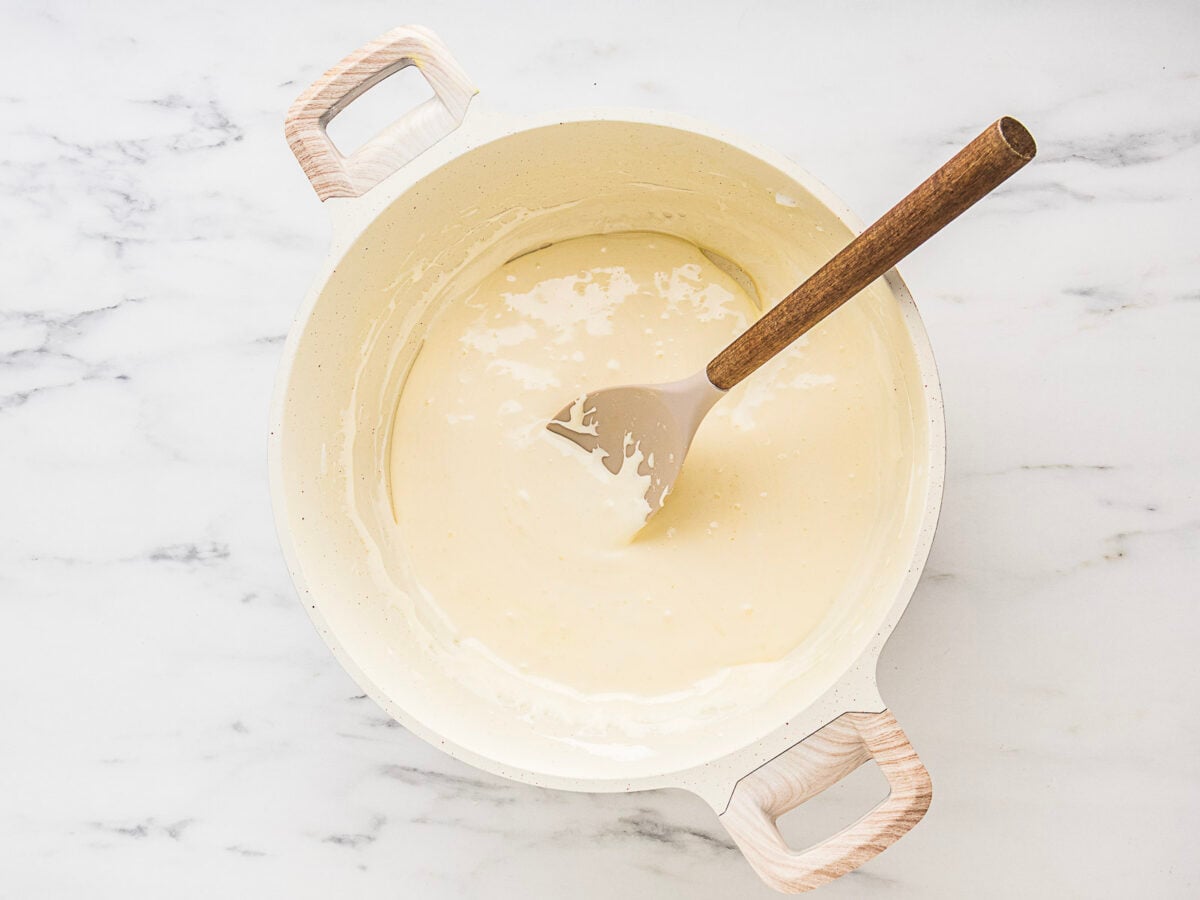 A white pot filled with a smooth, creamy sauce being stirred with a spatula on a marble surface.