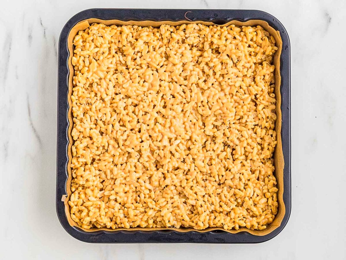 A square baking pan filled with golden rice krispie treats, lined with parchment paper, on a white marble surface.