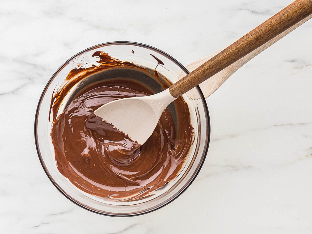 A glass bowl of melted chocolate being stirred with a wooden spoon on a white marble surface.