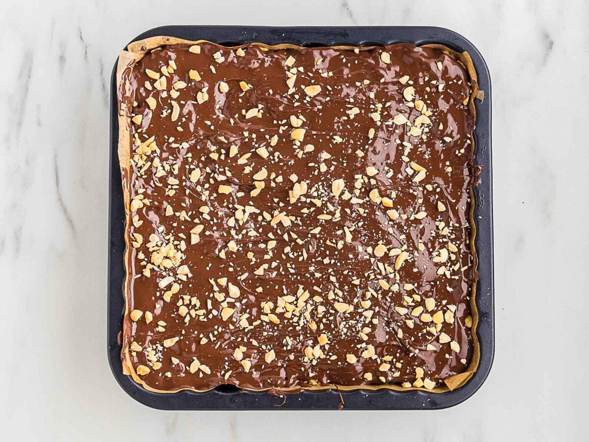 A square baking pan filled with a dessert topped with a layer of chocolate and sprinkled with chopped nuts, set on a white marble surface.