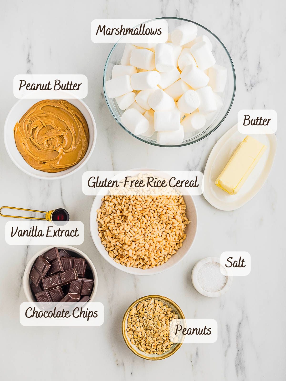 Bowls and plates containing marshmallows, peanut butter, butter, vanilla extract, chocolate chips, gluten-free rice cereal, salt, and peanuts arranged on a marble surface.