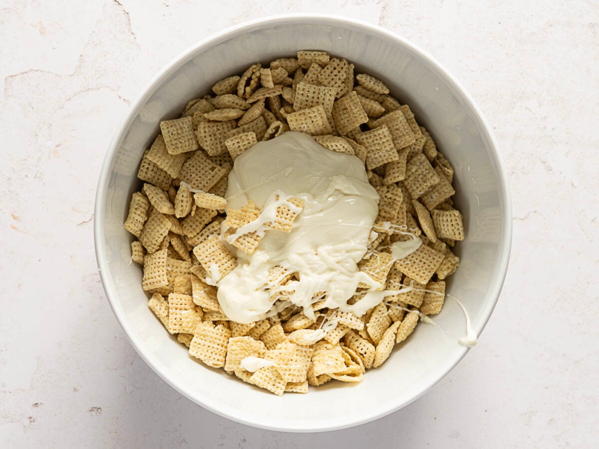 A white bowl filled with square cereal pieces, topped with melted white chocolate or yogurt, sits on a light-colored surface.