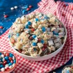 A bowl of patriotic snack mix with Chex cereal, red, white, and blue candies, sprinkles, and a red checkered cloth underneath.
