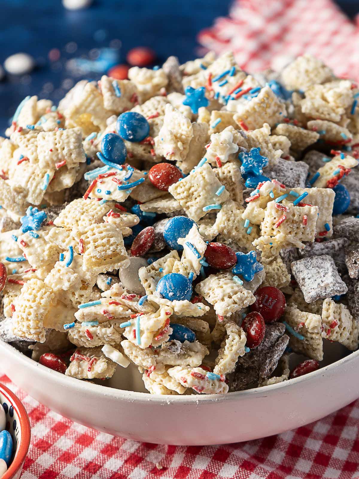A bowl of snack mix with Chex cereal, red and blue candy-coated chocolates, star-shaped candies, and sprinkles, set on a red checkered cloth.