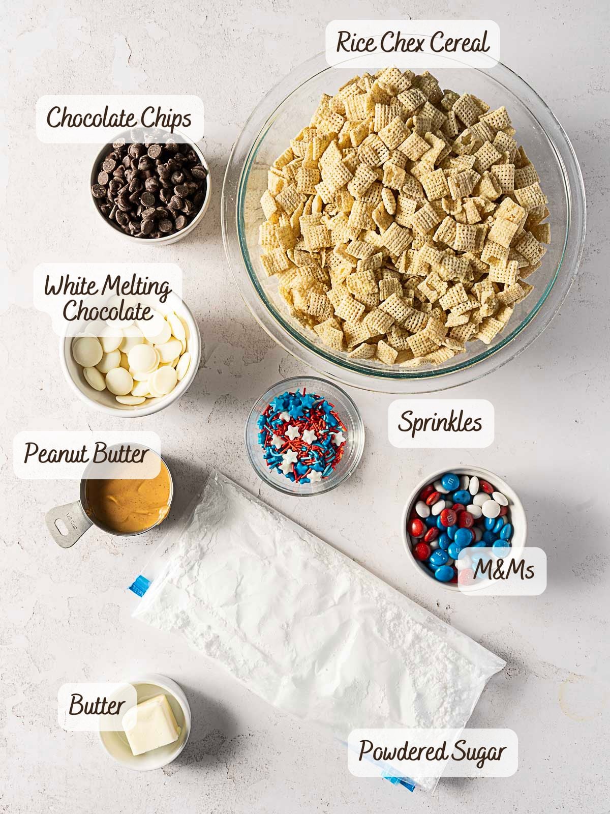 Bowls of Rice Chex cereal, chocolate chips, white melting chocolate, peanut butter, sprinkles, M&Ms, butter, and a bag of powdered sugar arranged on a white surface with labels.